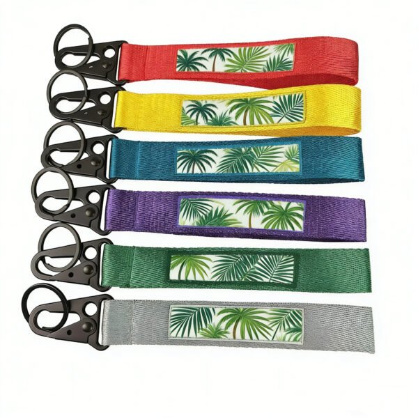 A flat-lay array of six vibrantly hued woven keychains, each emblazoned with verdant tropical palm prints atop a central white cartouche and crowned with matte black triangular lobster clasps, showcased against a pristine white void.