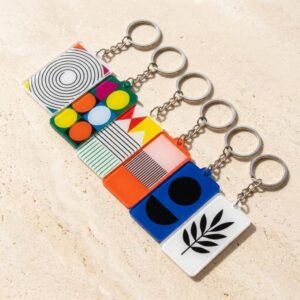 Custom acrylic keychain with square colorful pattern designs featuring concentric circles, color block patchwork, stripes, black-and-white geometric graphics, and botanical leaves, paired with a silver-tone metal chain and ring, on a beige sand and gravel background