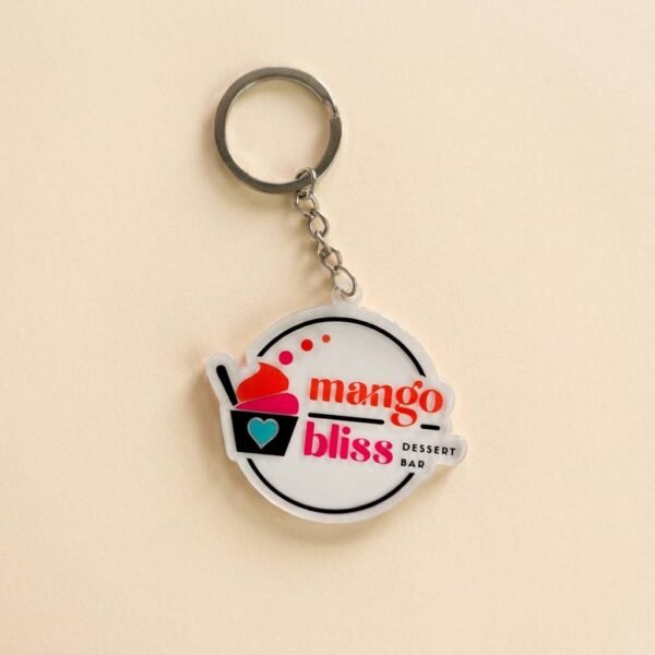 Round acrylic keychain featuring "mango bliss" lettering and a mango ice cream sundae illustration in a red, yellow, and pink contrasting color design, with a metal chain attached, displayed on a pure white background