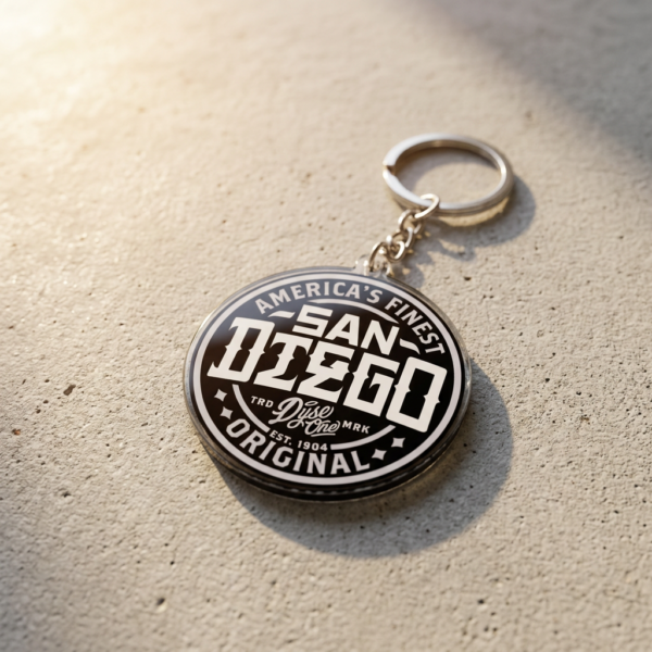 Custom acrylic keychain in a round design, featuring retro "SAN DIEGO" and "AMERICA'S FINEST" lettering in a black-and-white color scheme with transparent edges and a metal keyring at the top, placed on a concrete surface