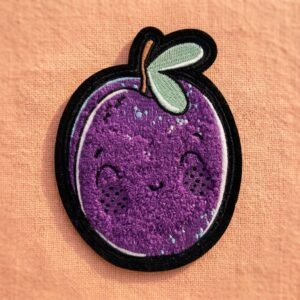 Custom chenille patch featuring purple cartoon fruits with smiley faces, green leaves, and brown stems, finished with black overlock edging, on a light orange linen background
