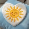 Custom chenille patch, heart-shaped white base fabric with a yellow smiling sun design at the center and pink blush accents, placed on light blue felt fabric