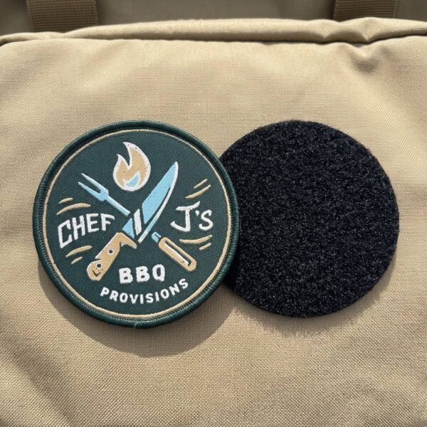 Custom embroidered Velcro patch featuring a Chef J's BBQ themed design with crossed knife and fork and flame elements, fully backed with Velcro, displayed against a khaki fabric background