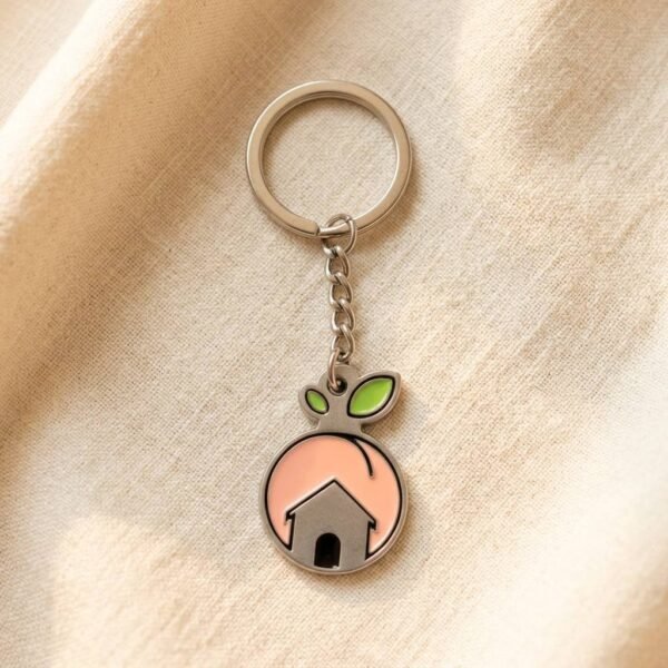 Custom round enamel keychain with silver house silhouette and green leaf accents on peach background, perfect for realtors & home gifts
