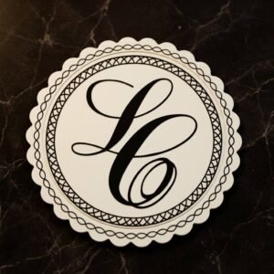 Custom PVC coaster with a cream-white round vintage scalloped design, featuring a black artistic monogram letter "L" at the center framed by a double-layer geometric woven border, placed on a dark marble textured background