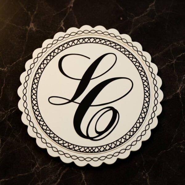 Custom PVC coaster with a cream-white round vintage scalloped design, featuring a black artistic monogram letter "L" at the center framed by a double-layer geometric woven border, placed on a dark marble textured background