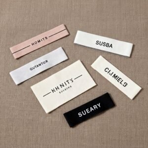 Custom Premium Woven Garment Neck Label Minimalist Letter Design Main Label Woven Tag Multiple premium woven garment labels in various colors and layouts scattered on a linen fabric background, featuring minimalist lettering designs including pink "HOMITS", white "SUSBA" and "GUTANTUR", beige "KN NITY", and black "SUEARY", showcasing fine weaving craftsmanship