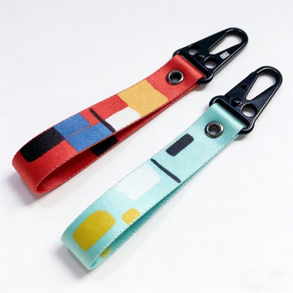 Custom webbing keychain featuring two artistic geometric print styles (Mondrian-inspired red, blue, yellow, black, and white design and mint green, yellow, and black abstract design), paired with a black metal lobster clasp, on a pure white background