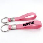 Custom pink silicone wristband keychain with black "MOVIE" lettering printed on the surface, featuring a metal connector and keyring — doubling as a wrist strap and keychain charm