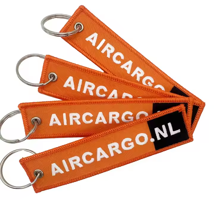 A set of four orange canvas embroidered keychains. Each features a white "AIRCARGO.NL" logo in bold text. The tags are attached to silver metal split rings via a woven loop. The design is minimalist, utilitarian, and highly visible, ideal for logistics branding.