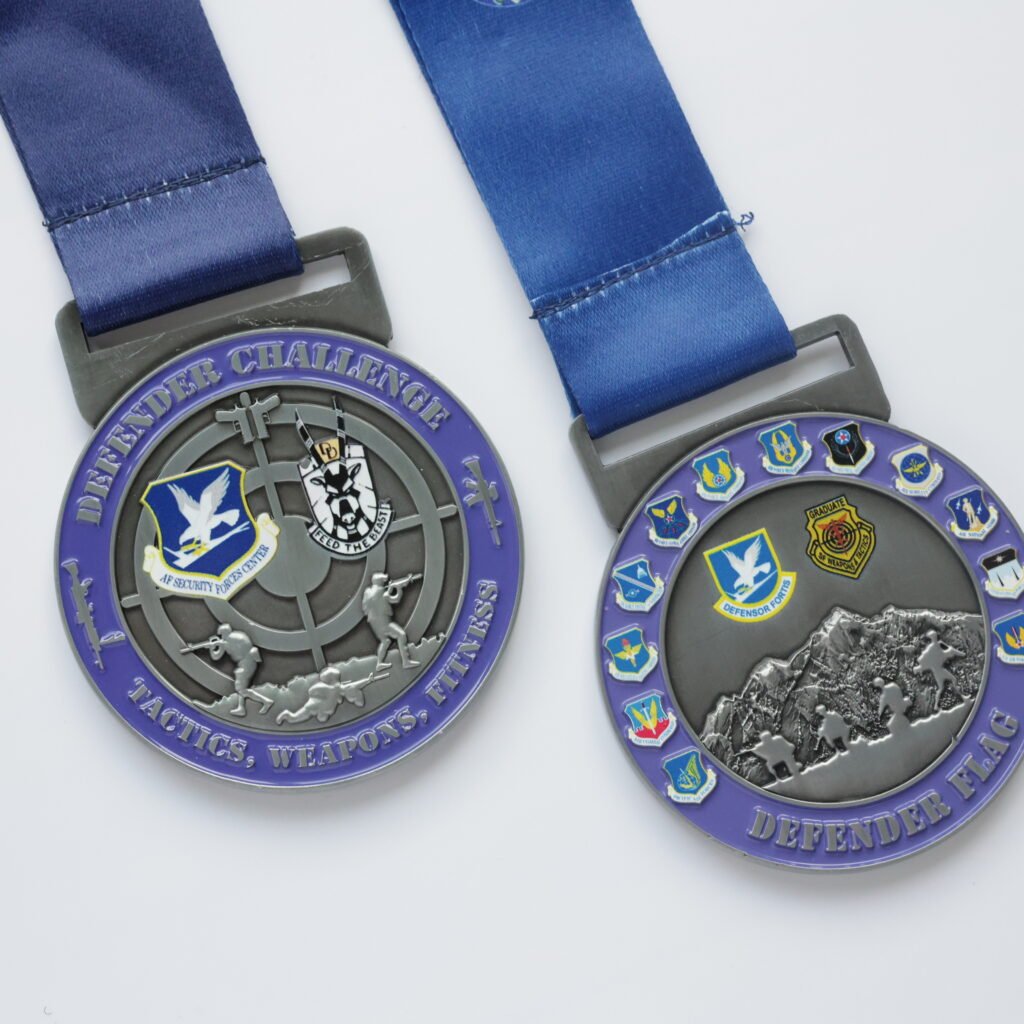 Two zinc alloy metal medals with a purple-and-silver dual-tone electroplating finish. The left medal features "DEFENDER CHALLENGE" lettering, a tactical figure running with a rifle, and an "AF SECURITY FORCES CENTER" badge on the front; the right medal features "DEFENDER FLAG" lettering, a snow mountain march embossed scene, and multiple commemorative badges, both paired with blue polyester ribbons, displayed on a pure white background