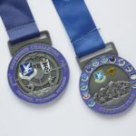 Two zinc alloy metal medals with a purple-and-silver dual-tone electroplating finish. The left medal features "DEFENDER CHALLENGE" lettering, a tactical figure running with a rifle, and an "AF SECURITY FORCES CENTER" badge on the front; the right medal features "DEFENDER FLAG" lettering, a snow mountain march embossed scene, and multiple commemorative badges, both paired with blue polyester ribbons, displayed on a pure white background