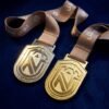 Custom metal medal in gold and silver dual versions, featuring a shield-shaped body with an engraved abstract "W" or "M" letter mark, paired with a brown woven ribbon, displayed on a dark blue velvet background — premium finish with refined luster