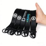 A set of custom black corporate webbing keychains featuring various company logos and text, including "DURABUILT," "NANO SERVICE," and a circular wave-pattern emblem. Each keychain is constructed from sturdy black nylon twill webbing with a robust silver metal triangle buckle and a D-ring, presenting a minimalist, professional, and durable aesthetic.
