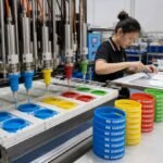 An industrial liquid silicone color infill machine automatically dispensing vibrant pigment into wristband molds, alongside neat stacks of finished blue, yellow, red, and green customized silicone wristbands during factory inspection.