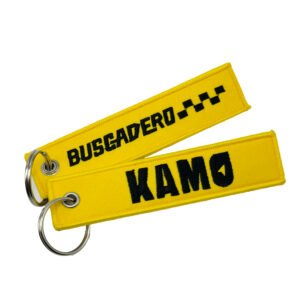 A yellow custom embroidered keychain. The front displays "BUSCADERO" text with a black and white checkered racing flag pattern, while the back shows bold uppercase letters "KAMO." It features high-density flat embroidery, a metal key ring, and neat edge stitching.