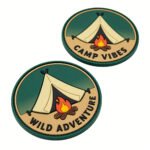Custom circular PVC soft rubber hook and loop patch with green camo edge, featuring a yellow camping tent and crackling campfire graphic, printed with "WILD ADVENTURE" and "CAMP VIBES" text; backed with heavy-duty nylon hook tape for attachment to outdoor backpacks, tactical vests or scout uniforms.