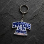 T-shirt shaped acrylic keychain printed with "TATTOOS WITH PASSION" and "BAYSIDE INK" text; features a silver metal ring and chain on a dark rock background.