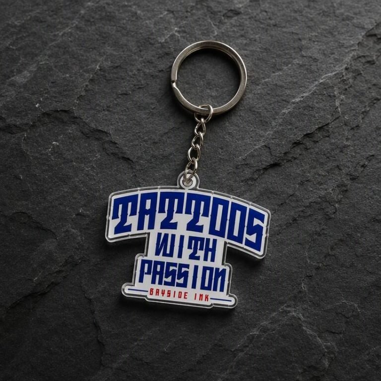 T-shirt shaped acrylic keychain printed with "TATTOOS WITH PASSION" and "BAYSIDE INK" text; features a silver metal ring and chain on a dark rock background.