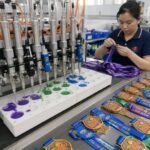 An automated multi-needle micro-injection machine dispensing liquid PVC into medal molds on a factory assembly line, with a technician quality-checking custom printed neck ribbons alongside a row of finished colorful 2D/3D PVC medals.